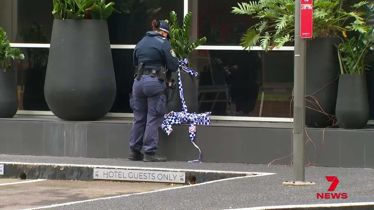 wide view of a police officer securing blue-and-white police tape at the Novotel entrance with 'hotel guests only' curb marking