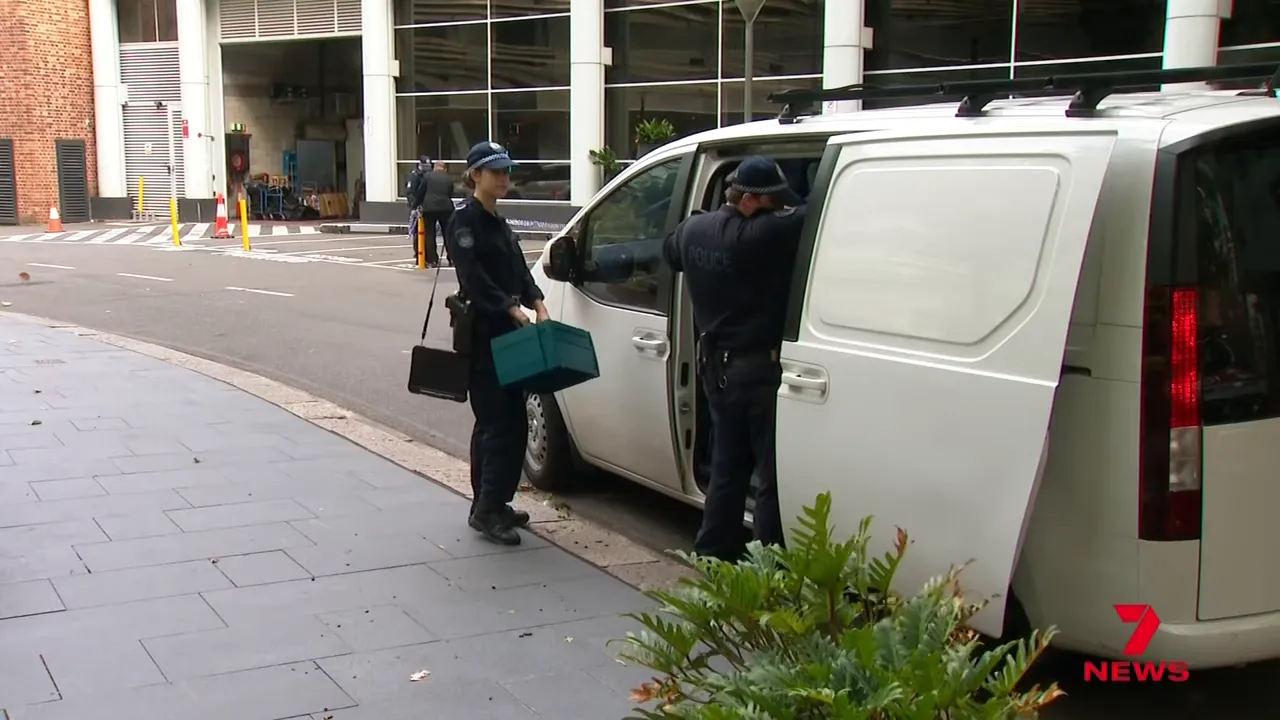 Two uniformed police officers beside a white van; one holds a teal evidence box while the other reaches into the vehicle at a hotel entrance
