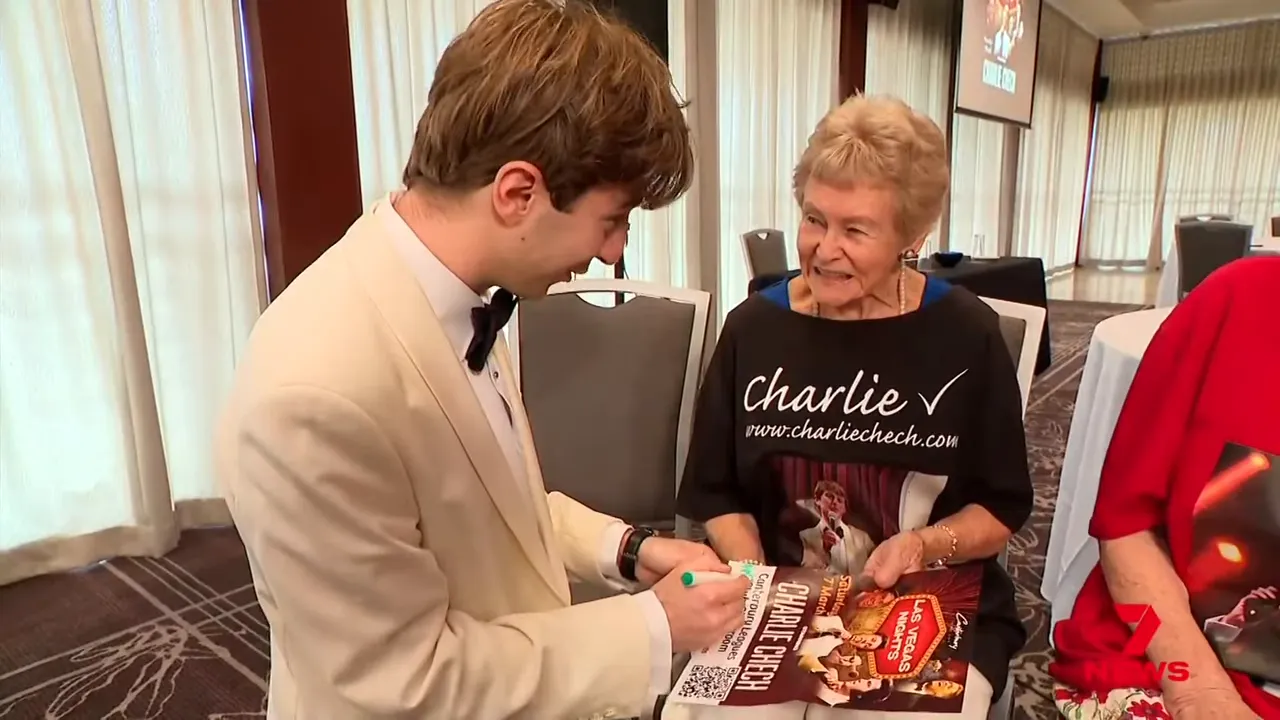 Performer in a white jacket signing a poster while an older fan looks on wearing a 'Charlie' T‑shirt