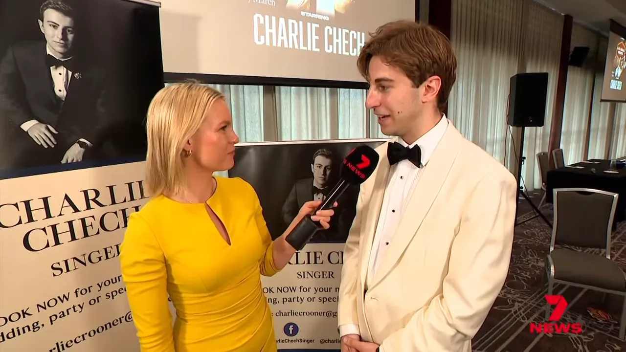 Wide shot of a presenter in yellow interviewing a singer in a white jacket with promotional posters and a conference room visible