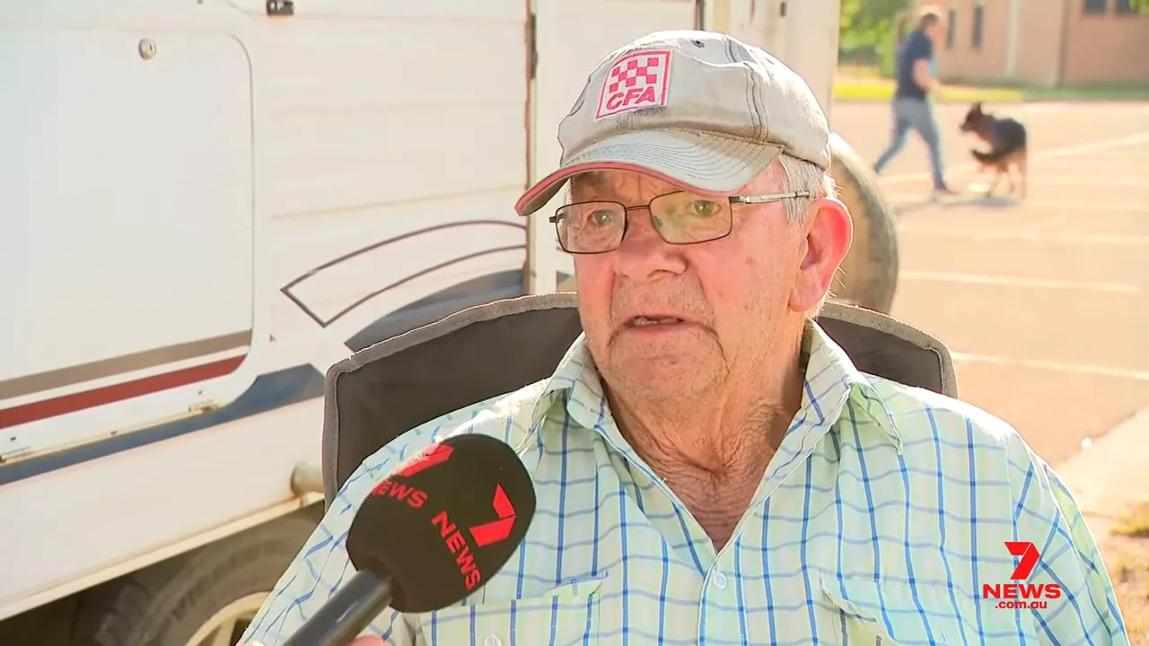 Elderly farmer being interviewed with a news microphone; a person and a dog are visible in the background, giving scene context.