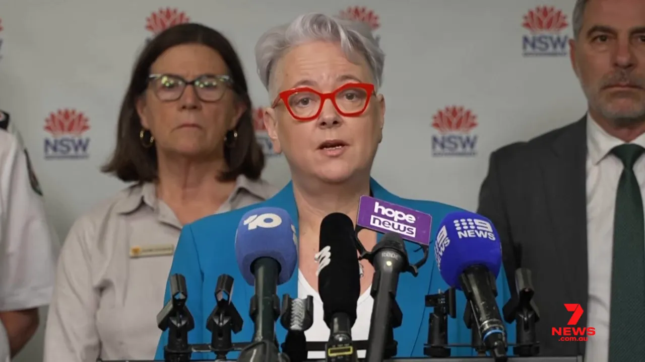 senior official speaking at a press briefing with microphones and colleagues behind