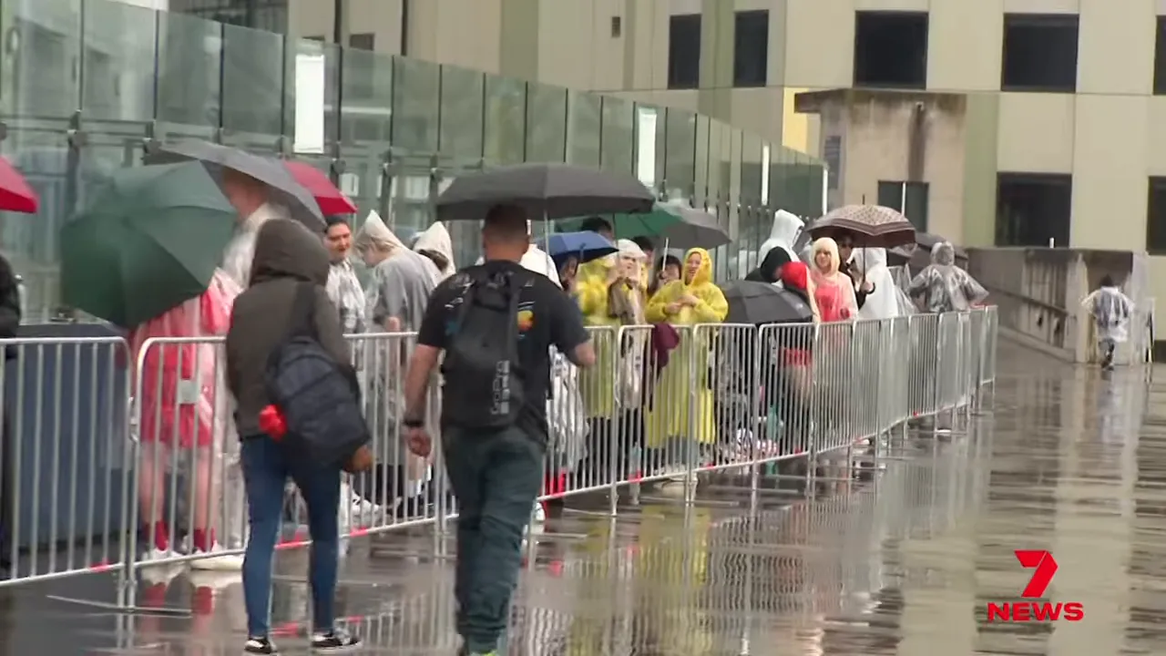 Long queue of fans in rain ponchos and under umbrellas behind metal barriers outside Marvel Stadium