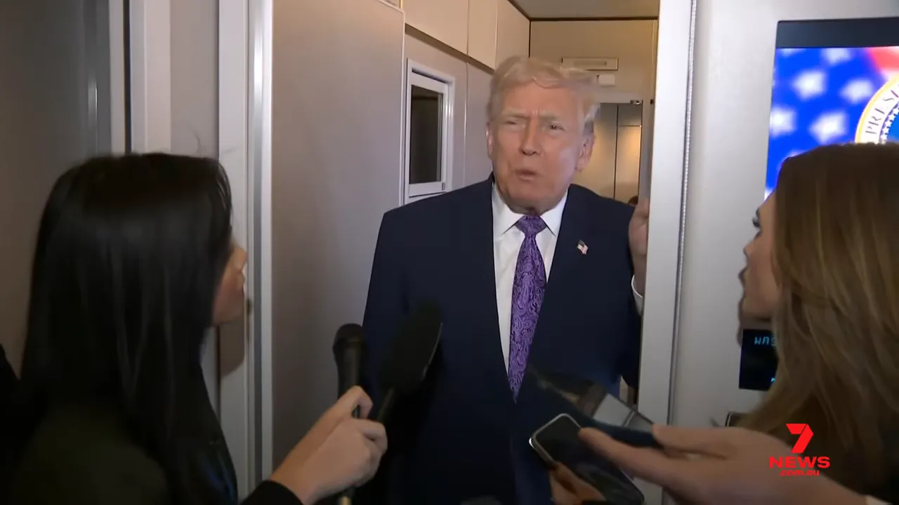 Senior official speaking from an aircraft doorway with reporters' microphones and phones visible, a screen with an emblem visible to the right.