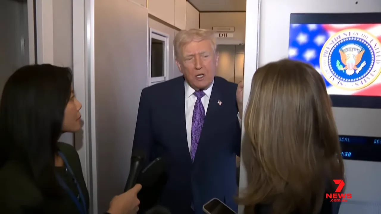 President speaking with reporters in an aircraft doorway with a presidential seal visible on a screen