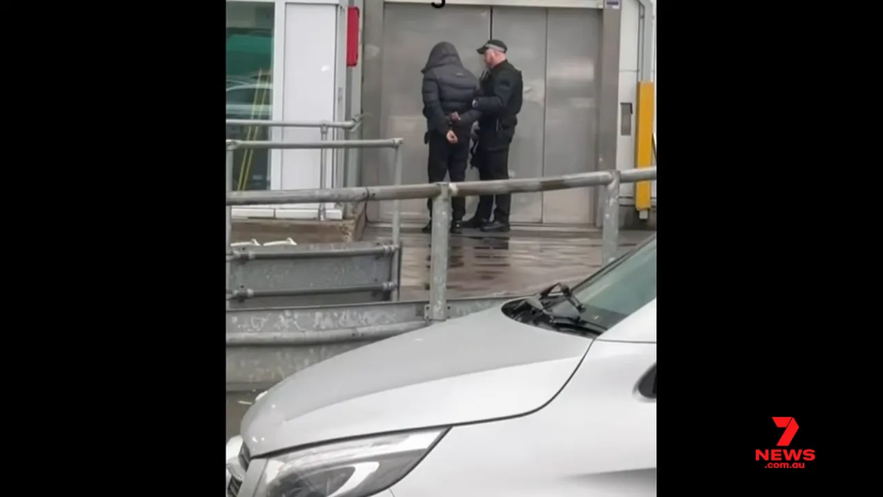 Hand‑cuffed person being escorted by a police officer outside elevator doors at Terminal Three