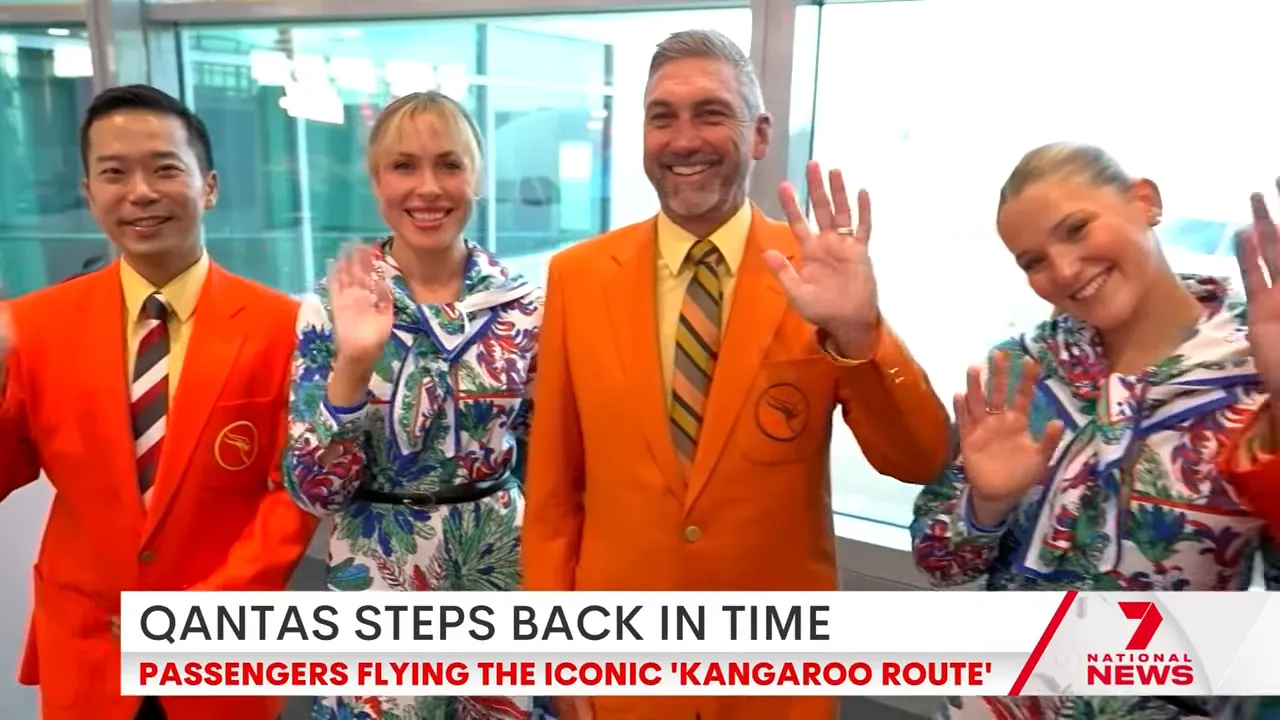Four Qantas crew members in bright orange jackets and patterned vintage dresses smiling and waving at the camera in an airport terminal