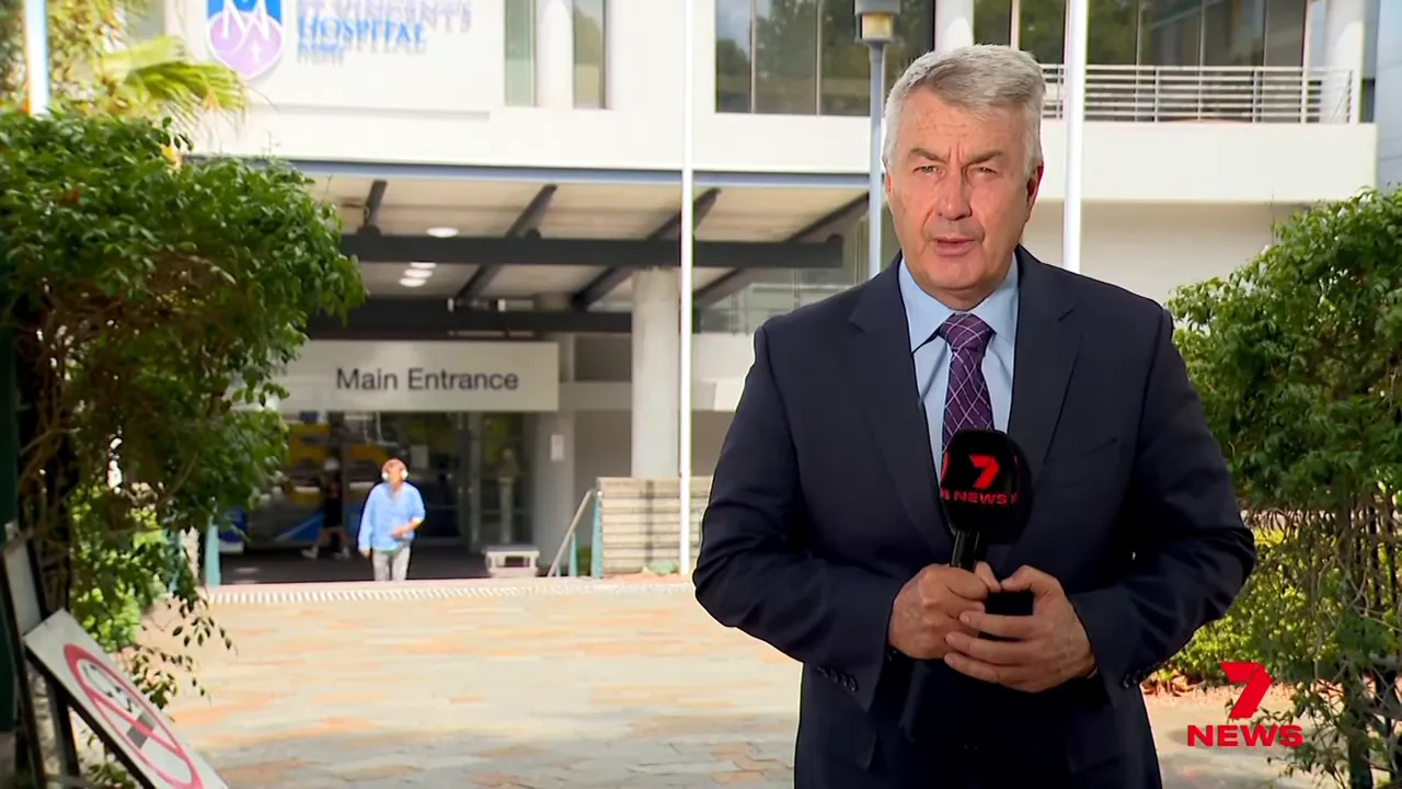 Television news reporter standing in front of a hospital main entrance with a person and signage visible in the background.