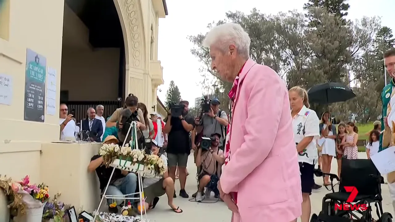 Elderly woman in a pink jacket standing before floral tributes and a wreath at the Bondi memorial with photographers and onlookers behind her