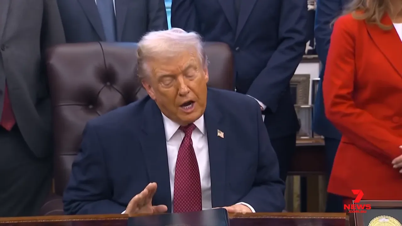 Man in navy suit at a desk with hand raised in a speaking gesture, people standing behind