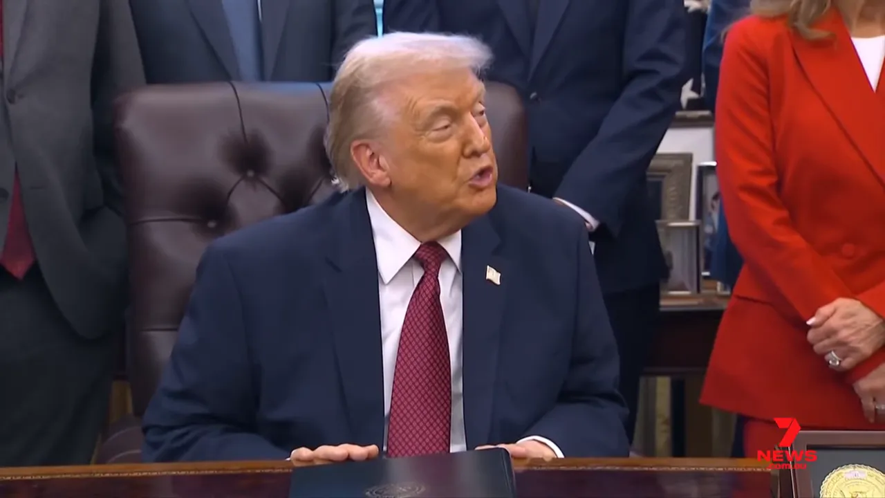 Profile view of a man in a dark suit speaking with both hands resting on a desk