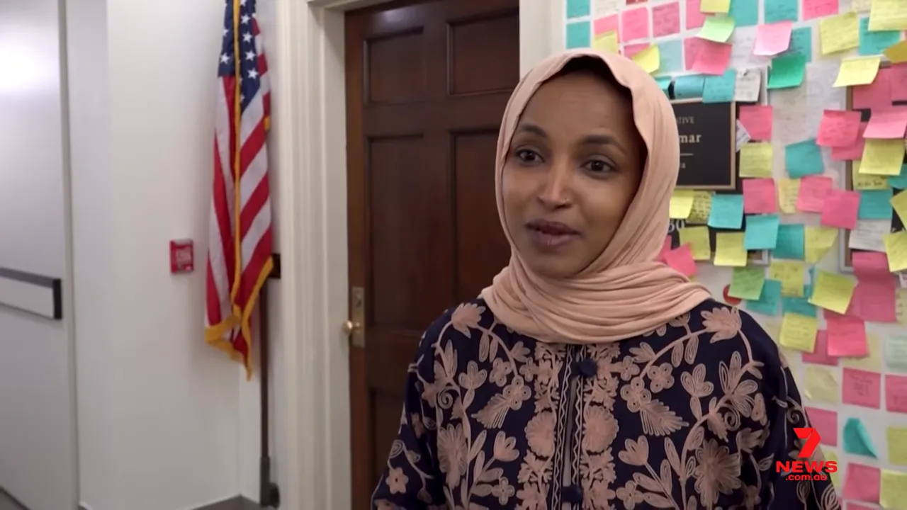 Ilhan Omar standing outside her office with a board of colorful sticky notes and a U.S. flag visible in the corridor.