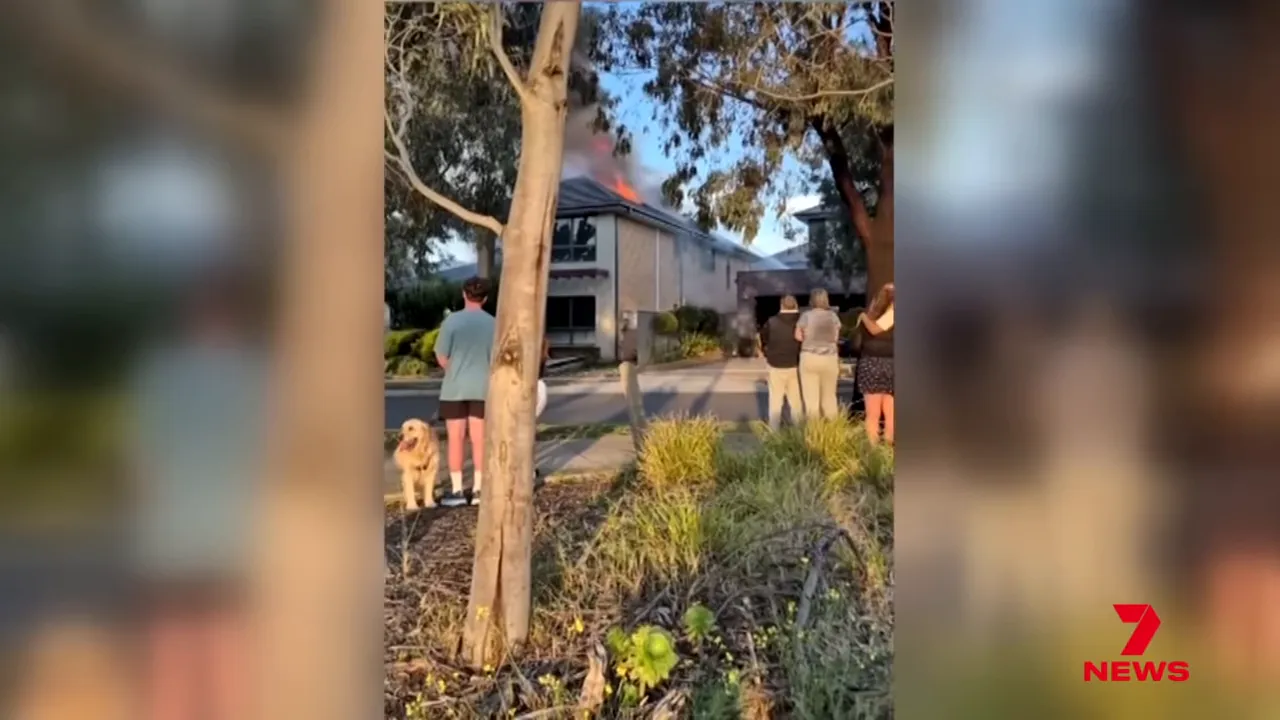 Bystanders watching a two-storey house with flames at the roof and smoke rising during a residential fire.