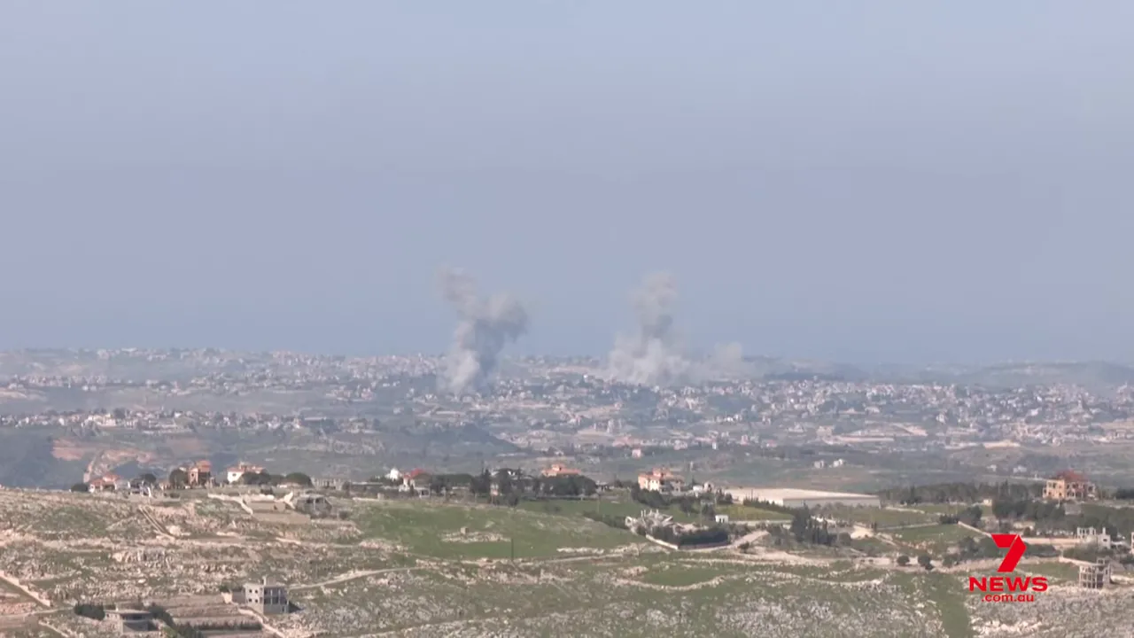 Daytime landscape with two large columns of smoke rising above a town after explosions; 7NEWS watermark.