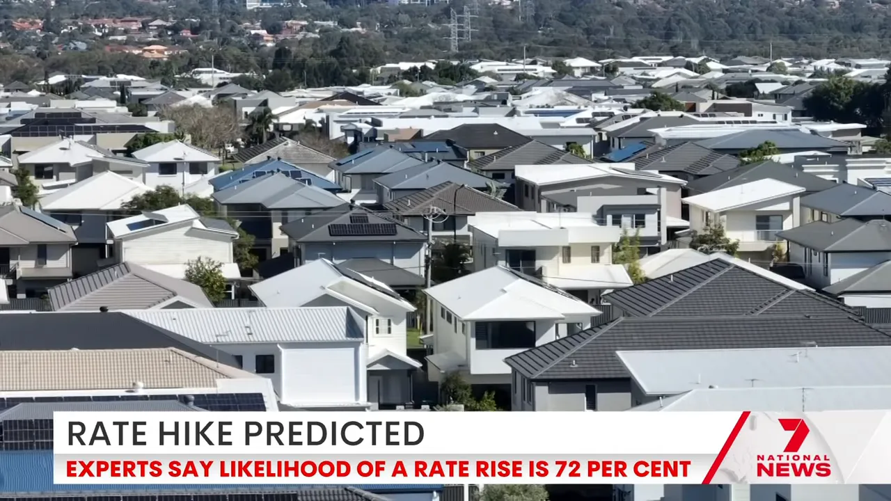 Aerial view of a dense suburban neighbourhood with many house rooftops and a news banner reading 'Rate Hike Predicted'.