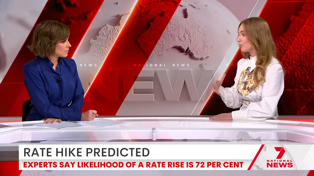 Wide shot of news desk interview with the 'RATE HIKE PREDICTED' banner across the bottom — anchor and guest in frame.