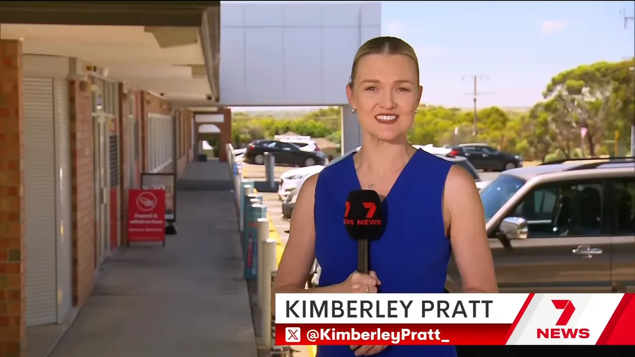 Wide shot of a TV reporter outside the Colonnades shopping centre showing the storefront walkway and carpark behind her.