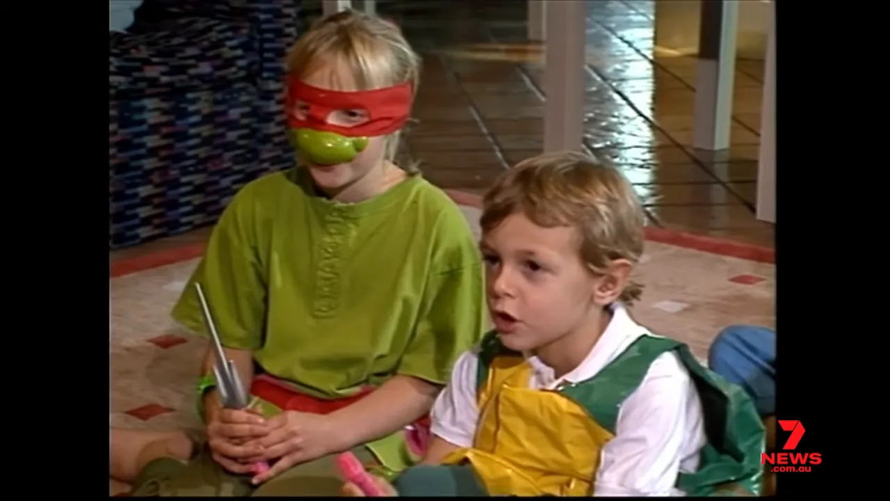 Two children in Teenage Mutant Ninja Turtles costume pieces holding toy weapons while role‑playing