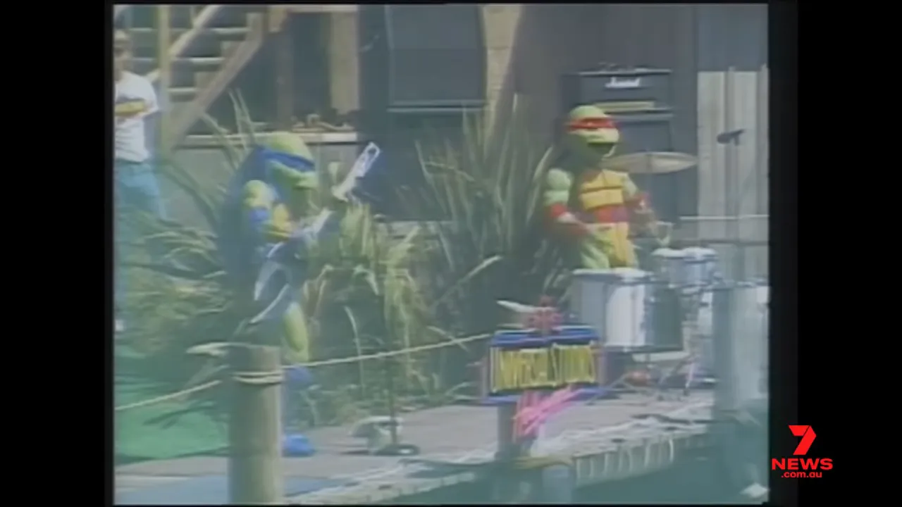 Two Teenage Mutant Ninja Turtles in costume performing on an outdoor stage with drums and signage