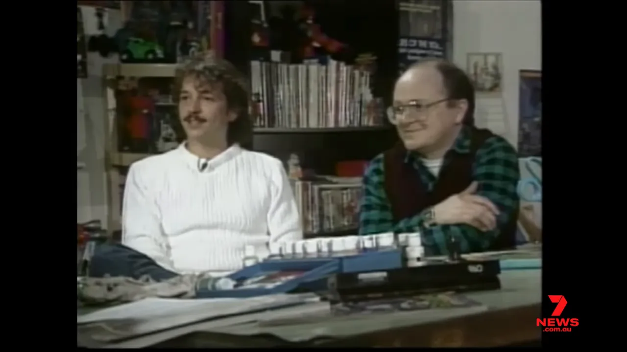 two men seated at a table during an interview with comics and toys on shelves behind them