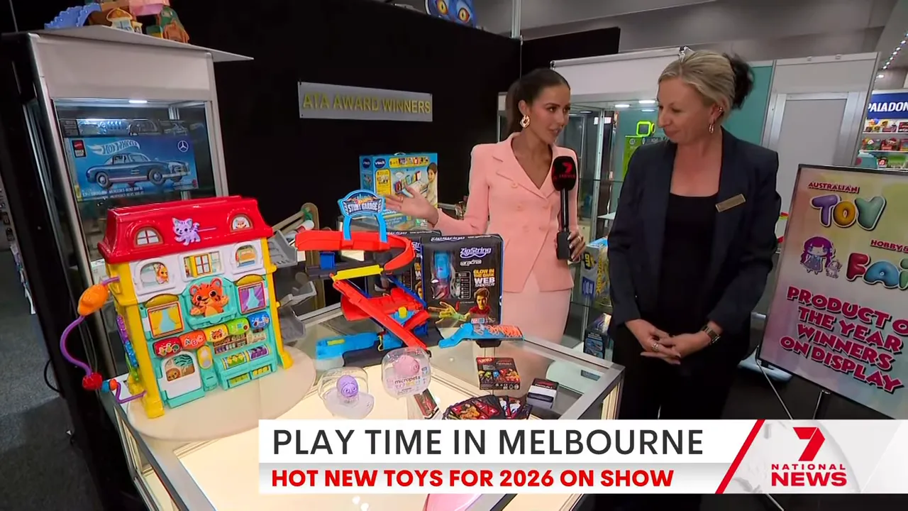 Display table at the toy fair featuring Micro Pets in clear pods, an activity peek-and-learn house and a red-and-blue stunt track, with presenters in the background