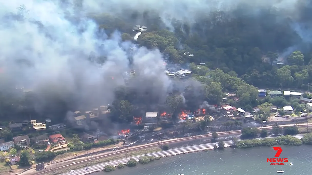 Wide aerial view of a coastline neighbourhood with houses burning, heavy smoke and a helicopter dropping water