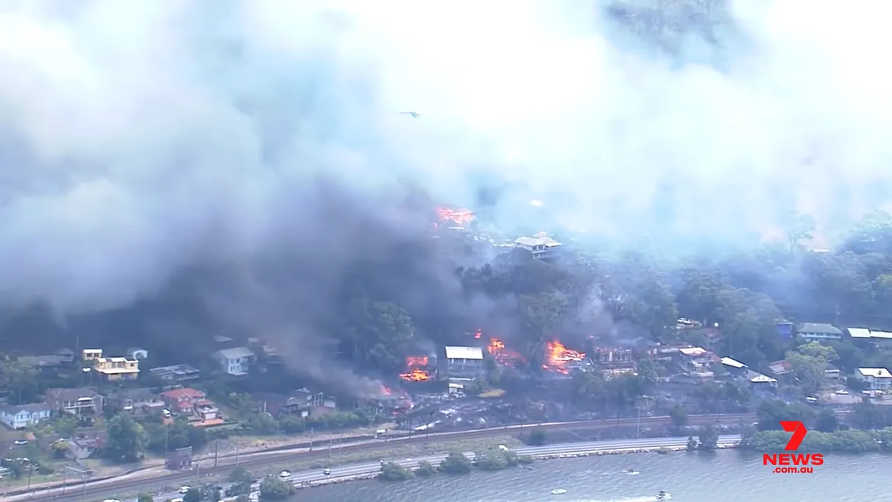 Wide aerial view of multiple houses on fire with heavy smoke across the neighbourhood and riverfront