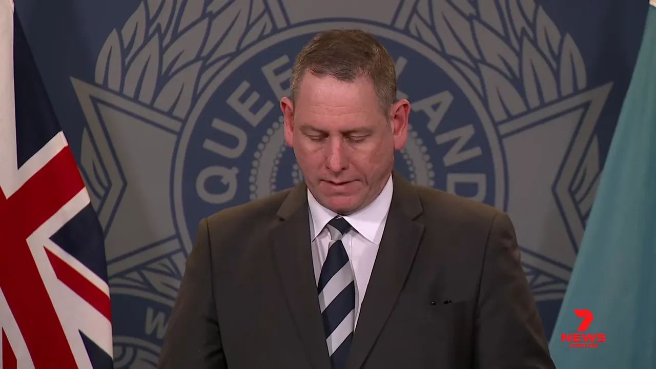 Queensland Police official at a press conference with police crest and flags in the background.
