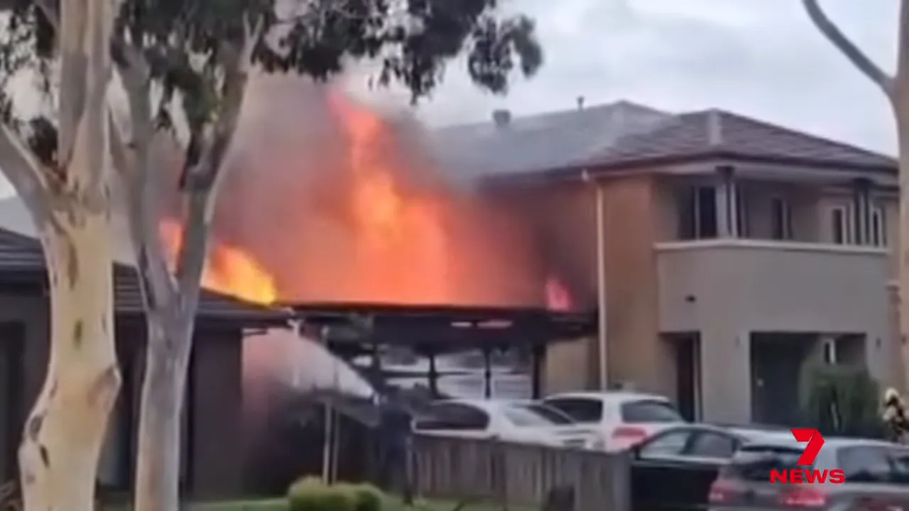 House garage on fire with large flames and a water spray aimed at the garage, cars parked outside.