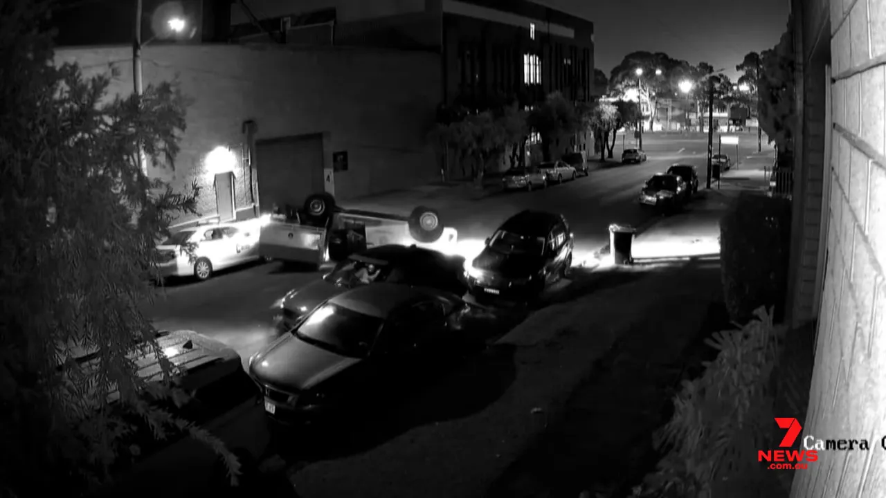 black-and-white CCTV image showing an overturned utility on its roof and damaged parked cars