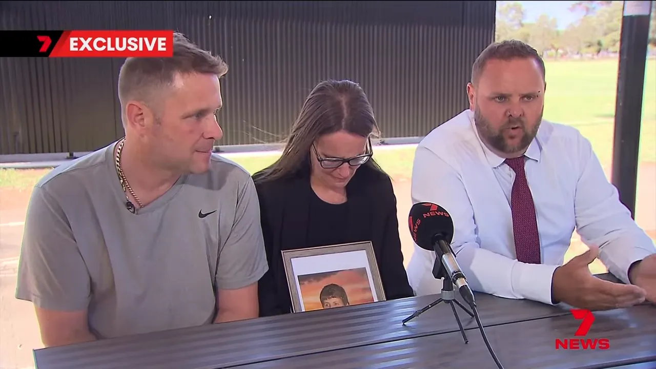 Three family members at a press interview holding a framed photo; one relative is speaking while another looks down, showing visible emotion.
