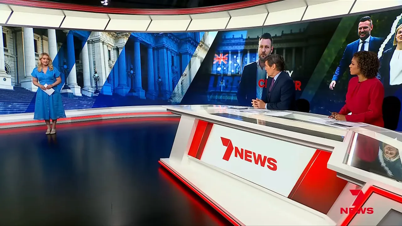 Television news studio with anchors at the desk and a large political graphic on the curved screen, showing live coverage and discussion.