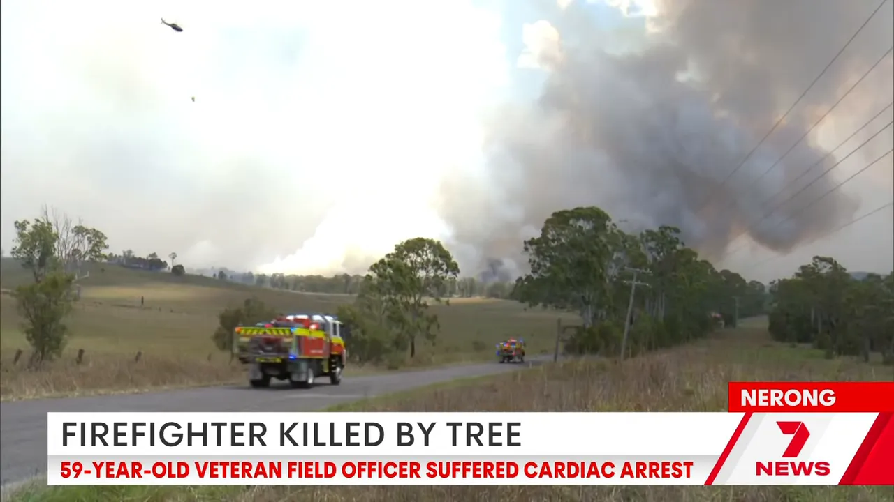 Fire trucks driving on a rural road toward a large smoke plume from a bushfire with a helicopter overhead.