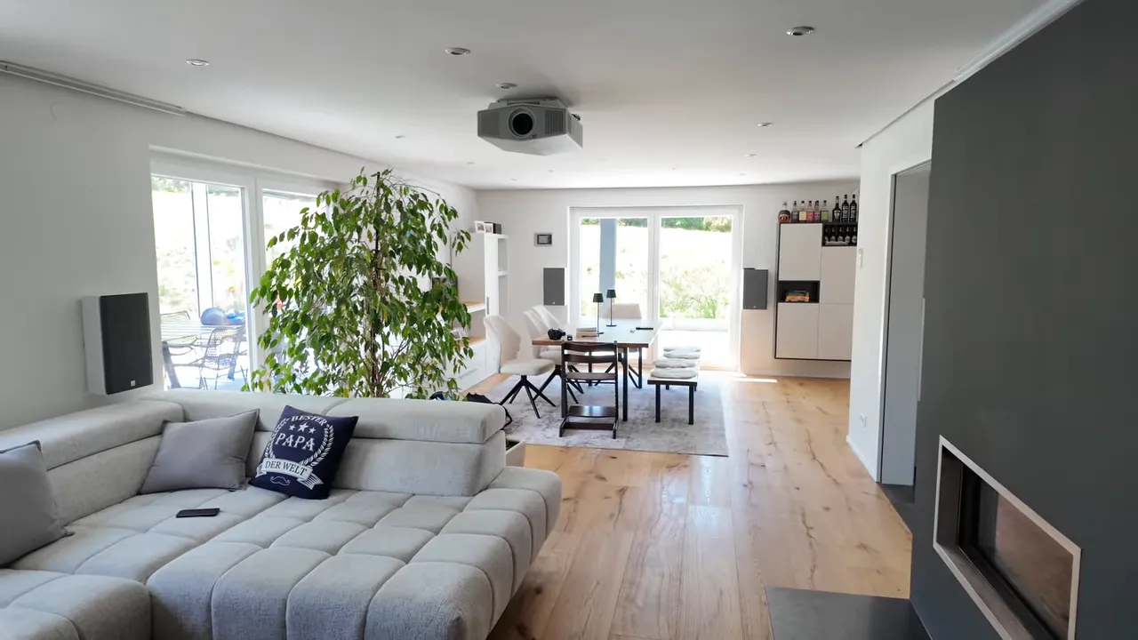 Offener Wohnraum mit grauem Sofa, Deckenprojektor, Essbereich und breiter Fensterfront