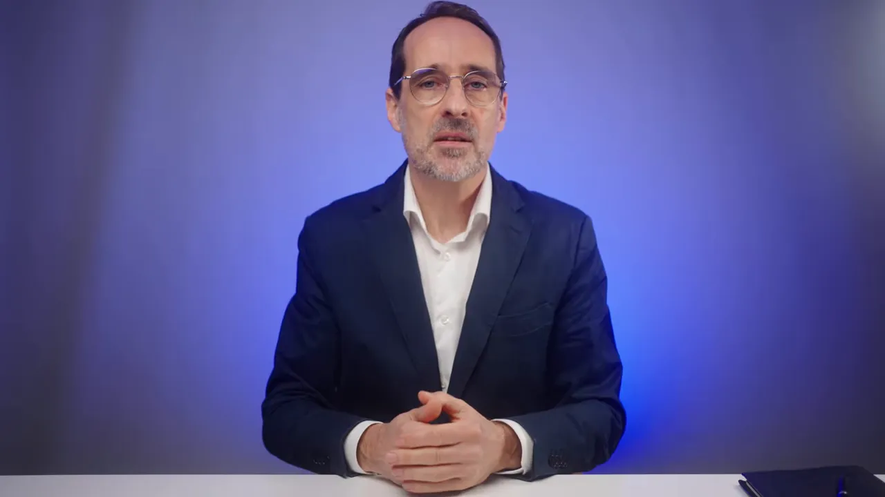 Referent vor blauem Hintergrund, ruhig sitzend mit gefalteten Händen auf dem Tisch