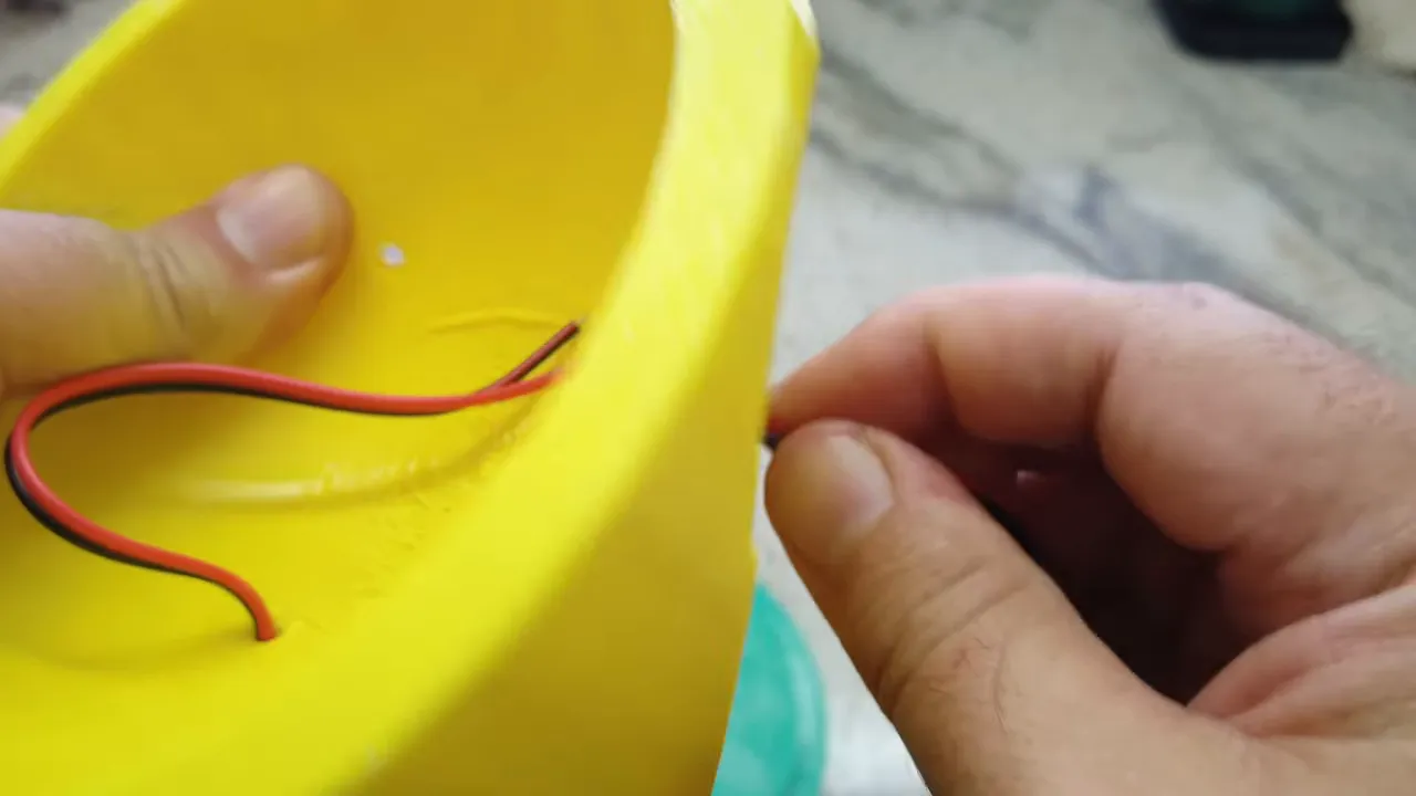 Red and black solar wires routed through holes in the interior of a yellow 3D-printed enclosure, hands positioning the wires.