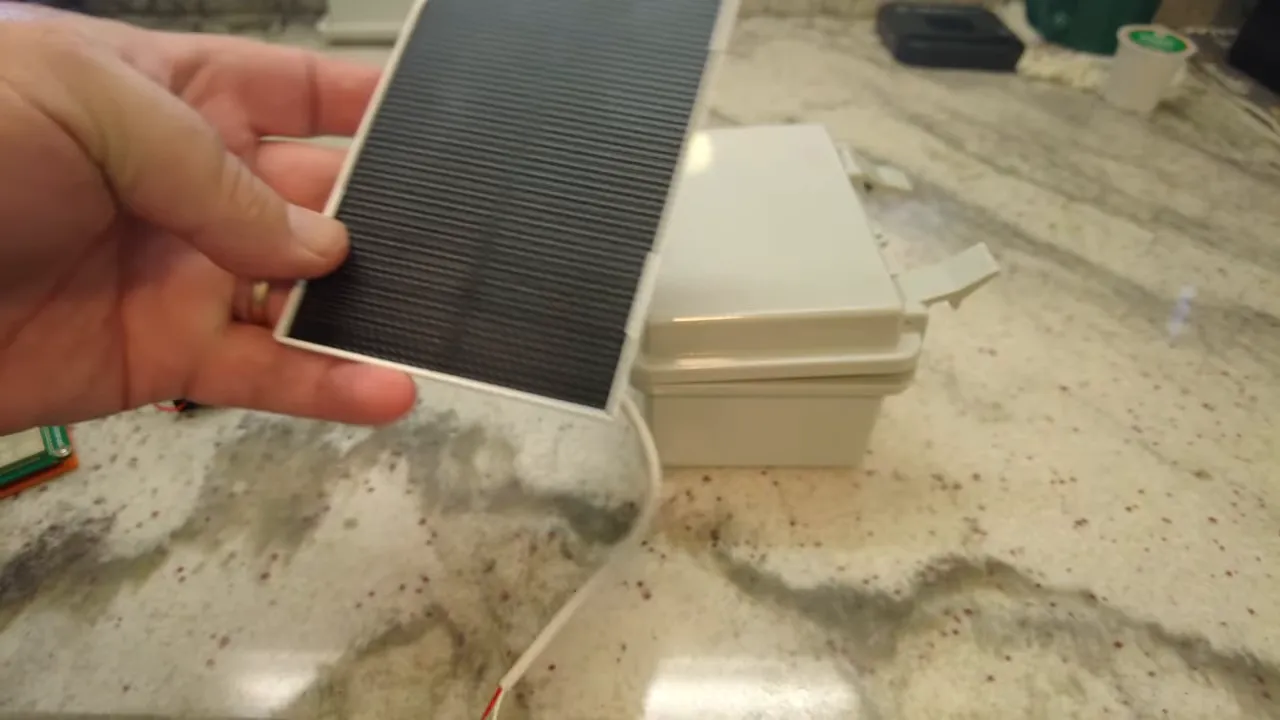 hand holding a small 3W solar panel with its cable next to a waterproof enclosure