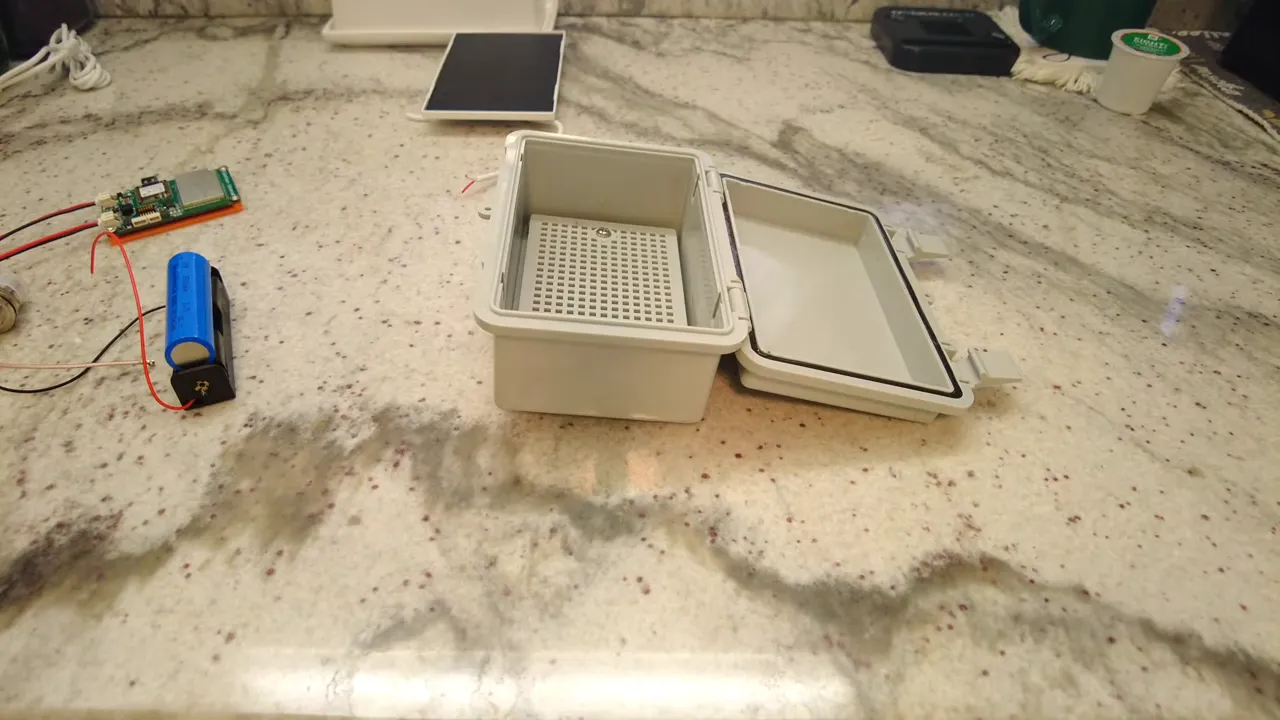 Top-down view of an open waterproof project enclosure with gasket, perforated interior tray, nearby battery, circuit board and small solar panel