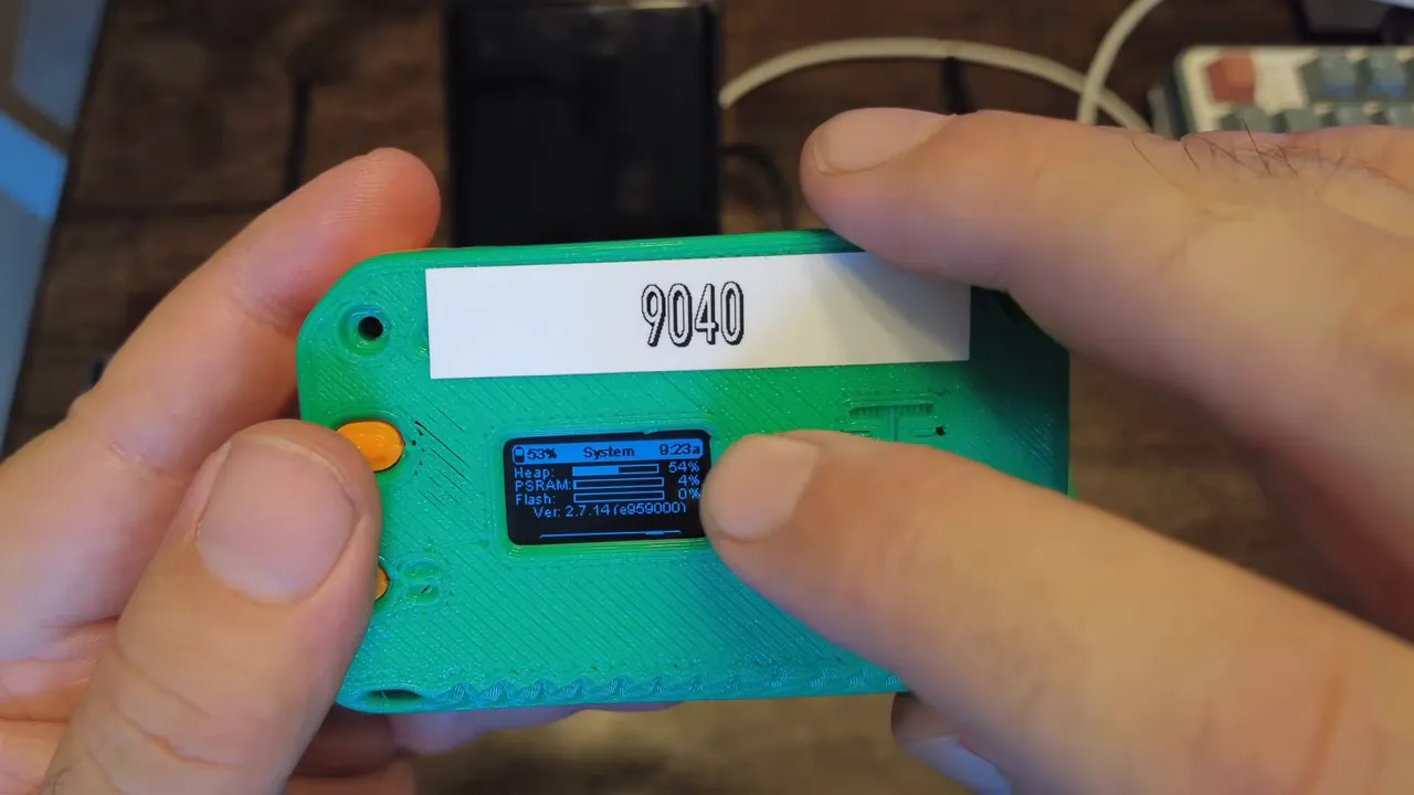 Centered view of a handheld green Meshtastic device with a finger pointing to the small system display that clearly shows PSRAM and heap values.