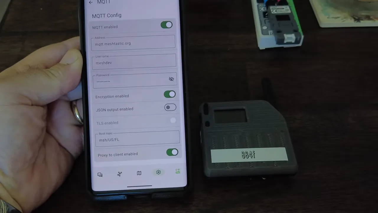 Meshtastic app MQTT configuration screen clearly showing address (mqtt.meshtastic.org), username, encryption and 'Proxy to client enabled' toggle with a Meshtastic LoRa node sitting next to the phone.
