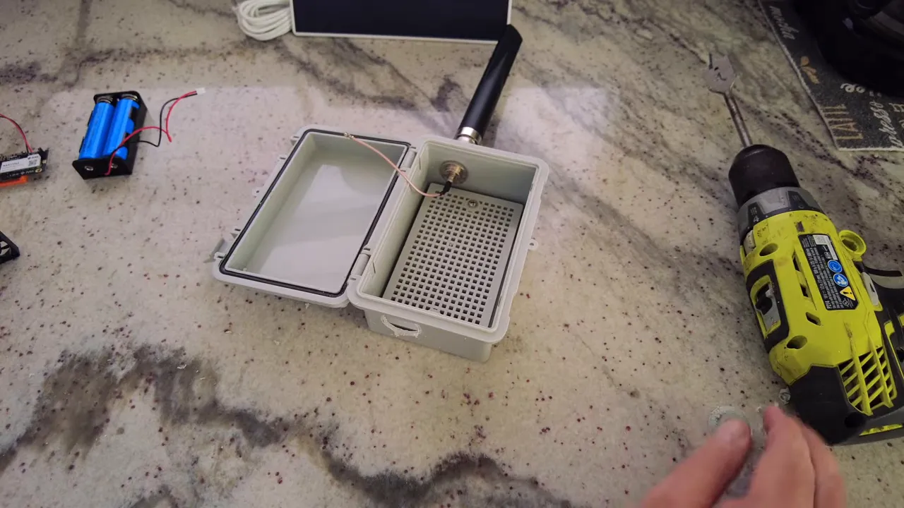 Open enclosure on a countertop with antenna feed and a drilled intake hole on the side, drill and battery pack nearby.