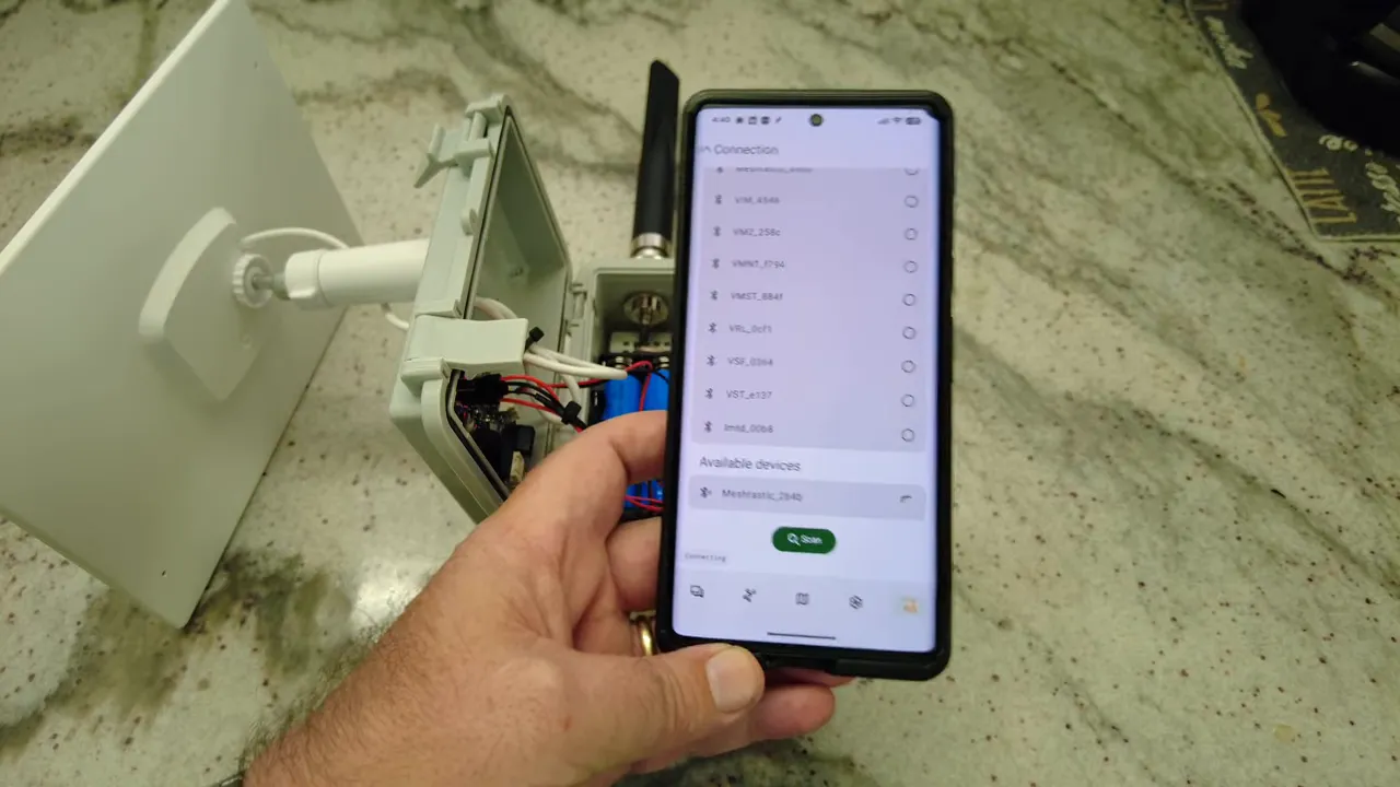 Smartphone displaying the Meshtastic app 'Available devices' section with 'Meshtastic_2b4b' highlighted; the open RAK solar node enclosure and antenna are visible on the counter.