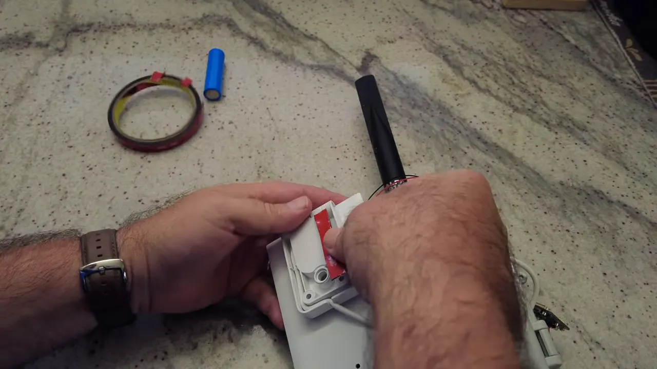 Hands applying double-sided tape to the enclosure mounting bracket next to the 915 MHz antenna