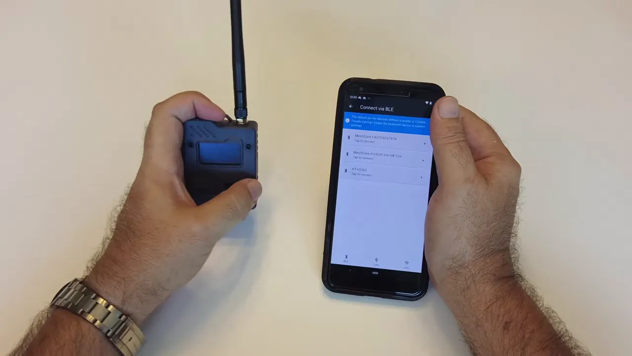 Hand holding the companion node beside a phone showing the MeshCore app pairing screen