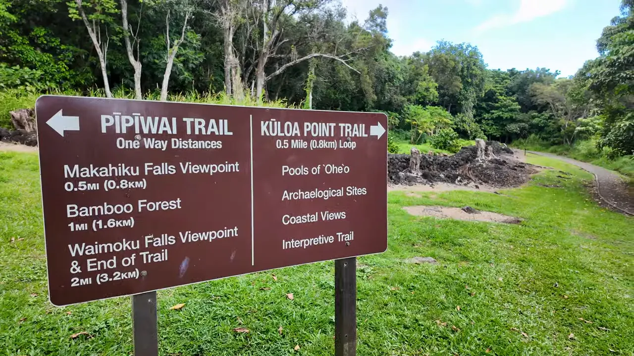 Pipiwai Trail sign showing distances to Makahiku Falls, Bamboo Forest, and Waimoku Falls beside the trailhead and greenery.