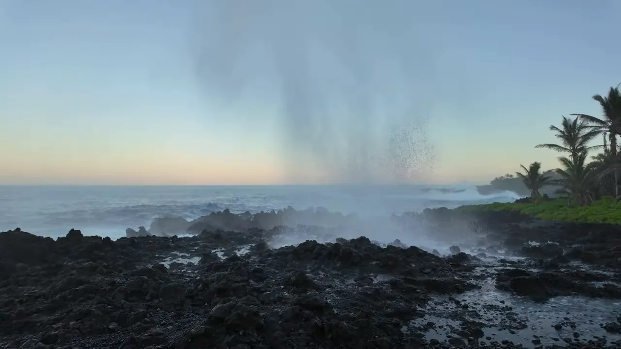 Ocean blowhole spouting water over dark lava rocks along the Waiʻānapanapa coastline at sunrise.