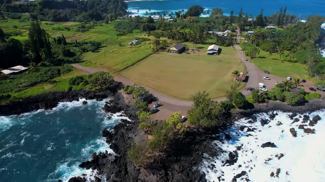 Aerial shot of Keʻanae Peninsula showing a grassy field, small buildings, parked cars and the lava-rock shoreline with waves.