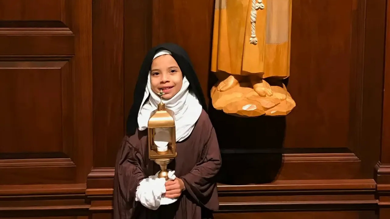 Child in a brown habit, white head covering, and black veil holding a gold lantern-shaped prop