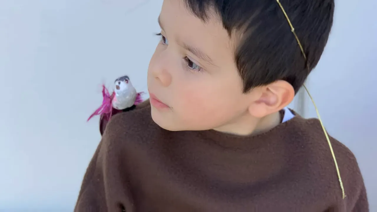 Child wearing a brown tunic and thin gold halo with an artificial bird on his shoulder