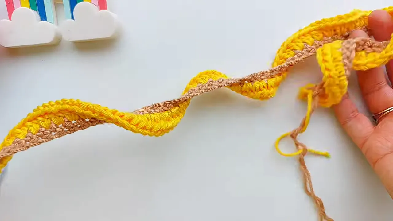 Beige crochet strip with a yellow outer row forming repeated waves on a white surface